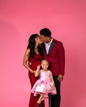 Pregnant couple sharing a kiss in red formal wear while their young daughter sits on stool in front on pink backdrop.