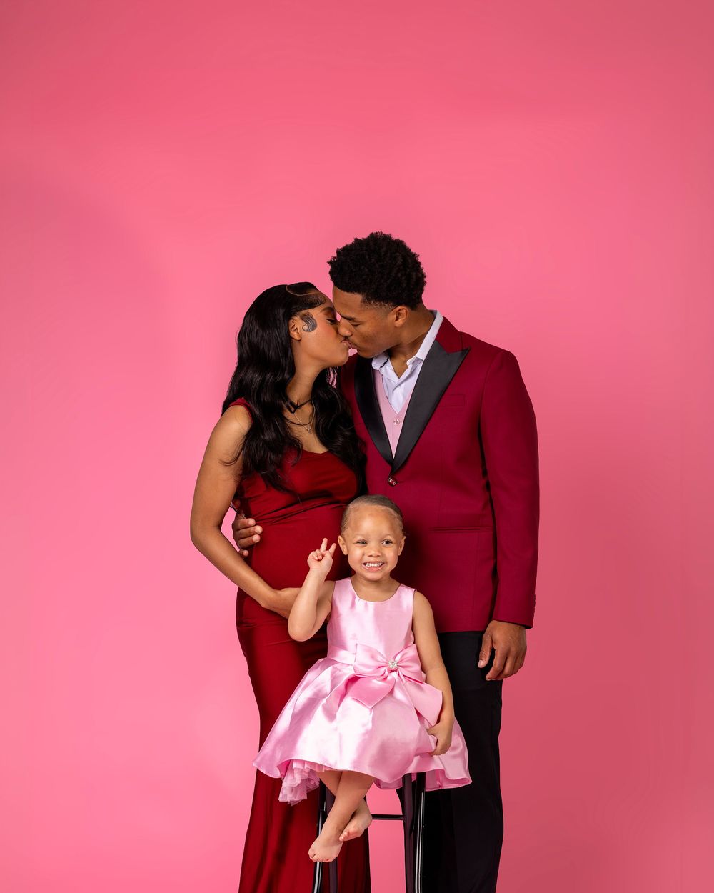 Pregnant couple sharing a kiss in red formal wear while their young daughter sits on stool in front on pink backdrop.