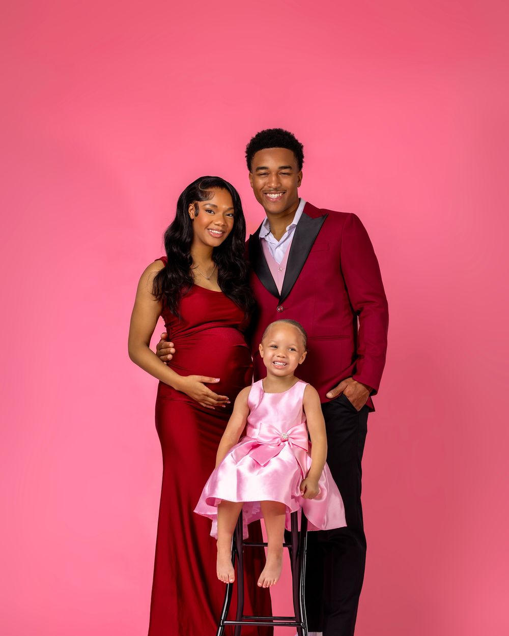 Expecting couple in matching red outfits with young daughter in pink dress posing for maternity photo on pink background.