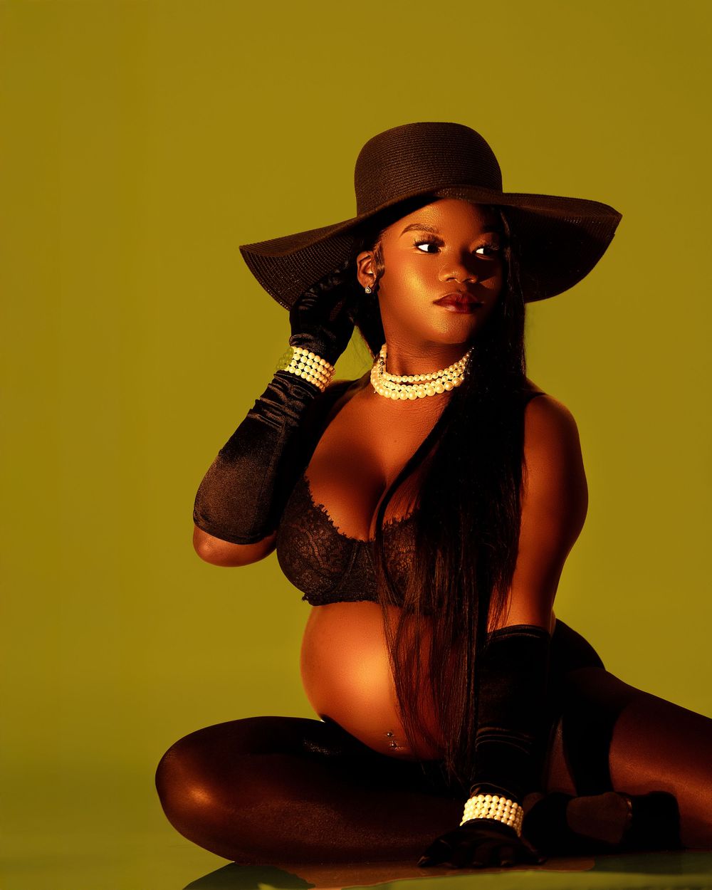 Expectant mother in black lace and wide-brim hat poses against vibrant yellow-green background.