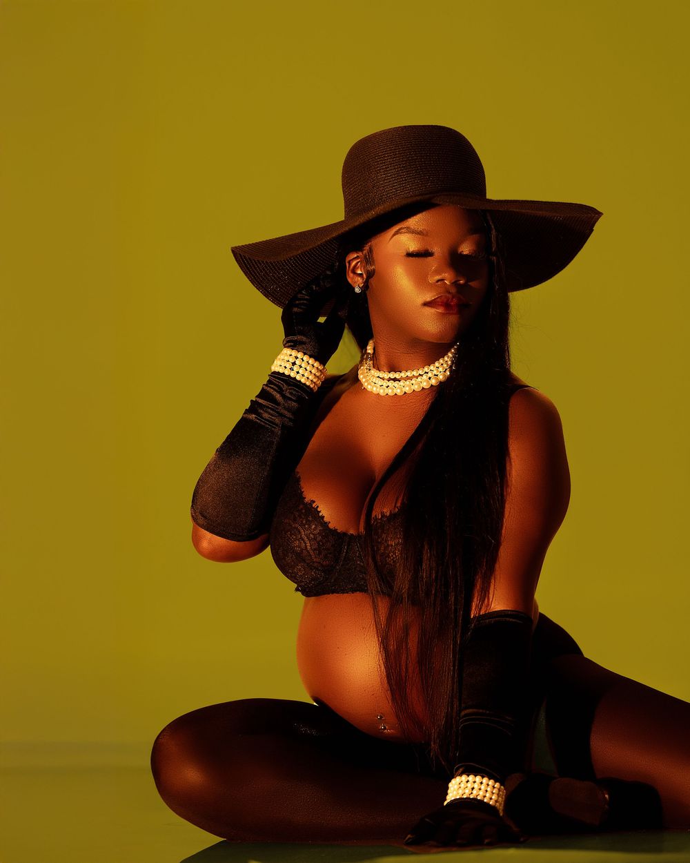 Pregnant woman in black lace bodysuit sits cross-legged wearing wide-brim hat and pearl jewelry.