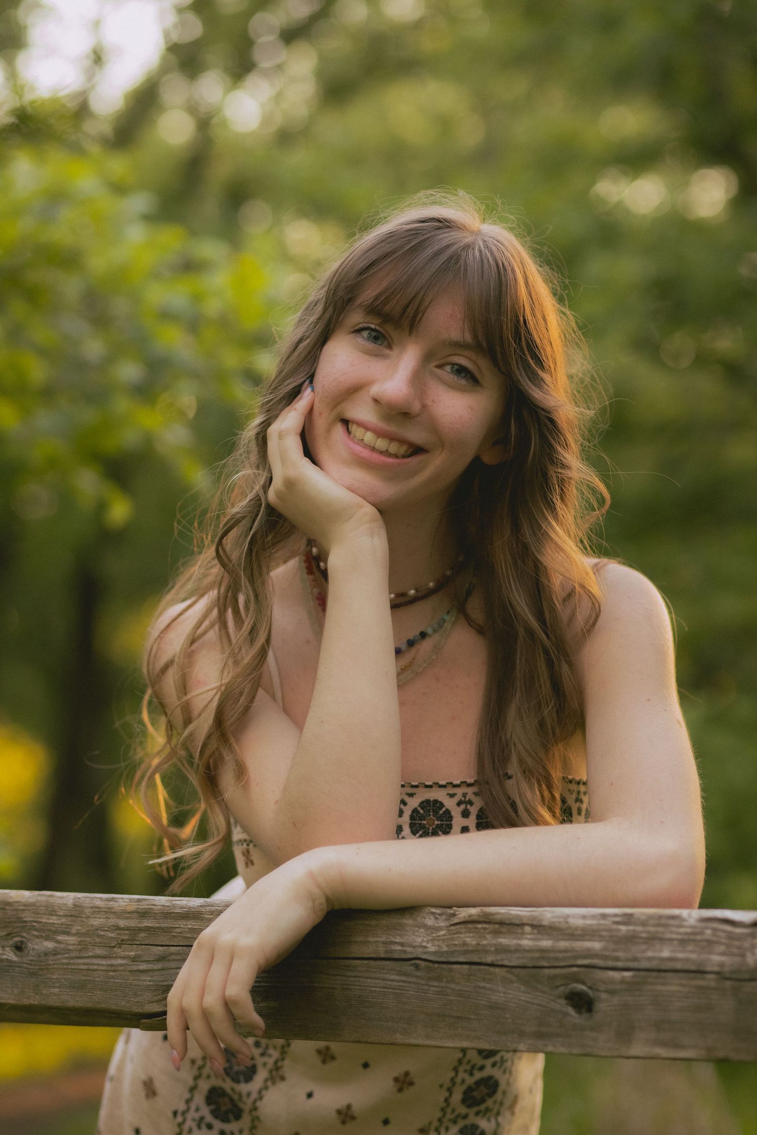 Smiling young woman with wavy hair leans on a wooden fence in a sunlit forest setting.
