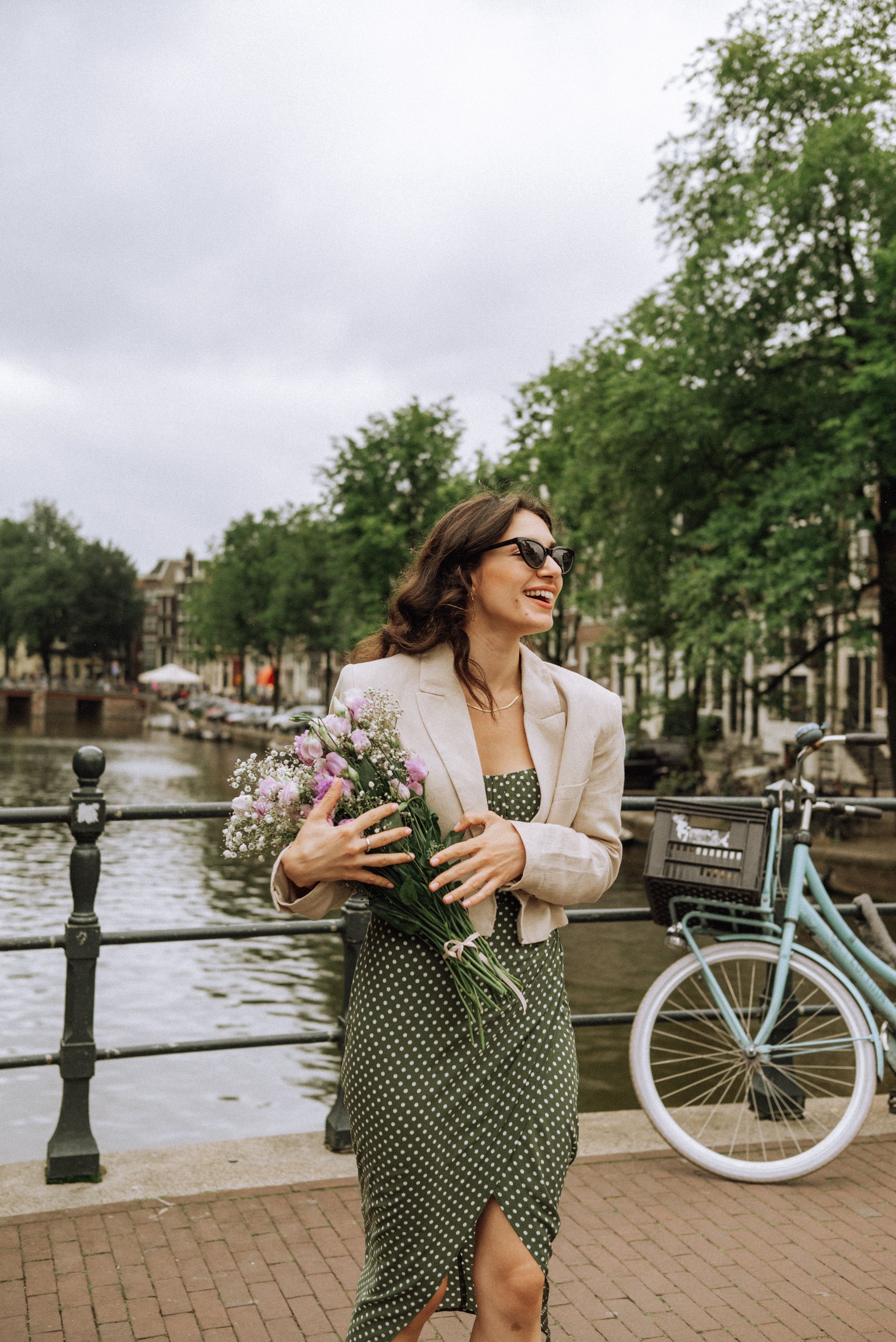 Woman in polka dot dress carrying flowers along Amsterdam canal with bicycle in background.