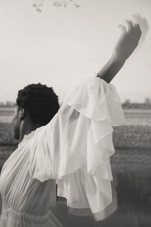 Artistic black and white portrait of a person in flowing white fabric against a rural backdrop.