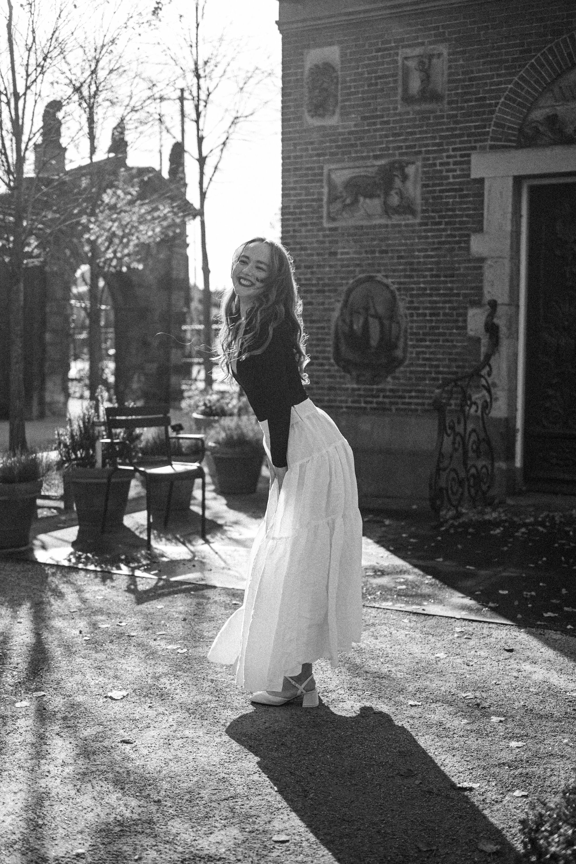 Black and white photo of person in flowing dress standing in sunlit garden setting.
