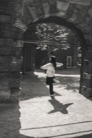 Figure walking under brick archways casting dramatic shadows on a sunlit path.