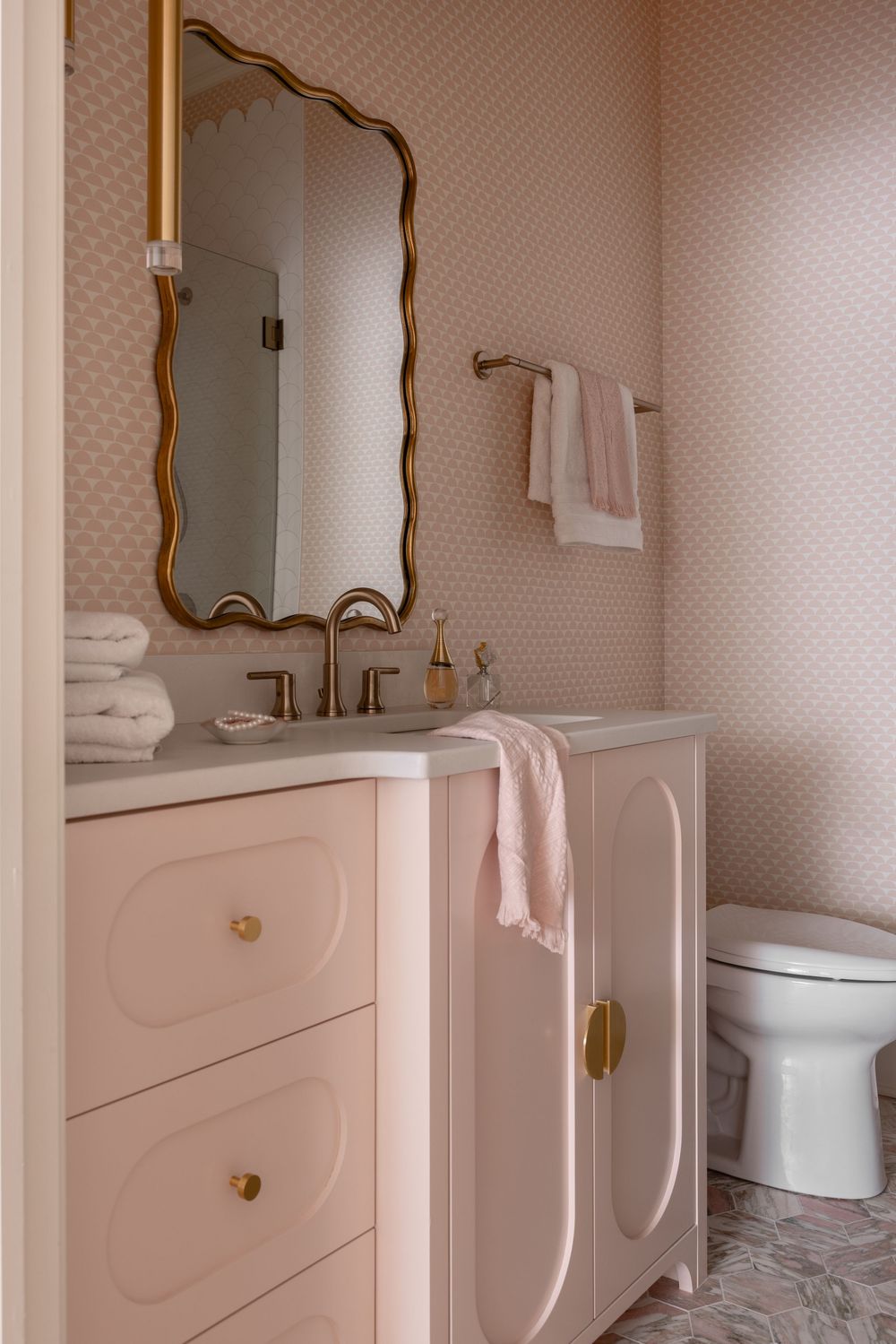 Pink bathroom vanity with gold hardware, wavy mirror, patterned wallpaper, and matching pink towels in elegant design.