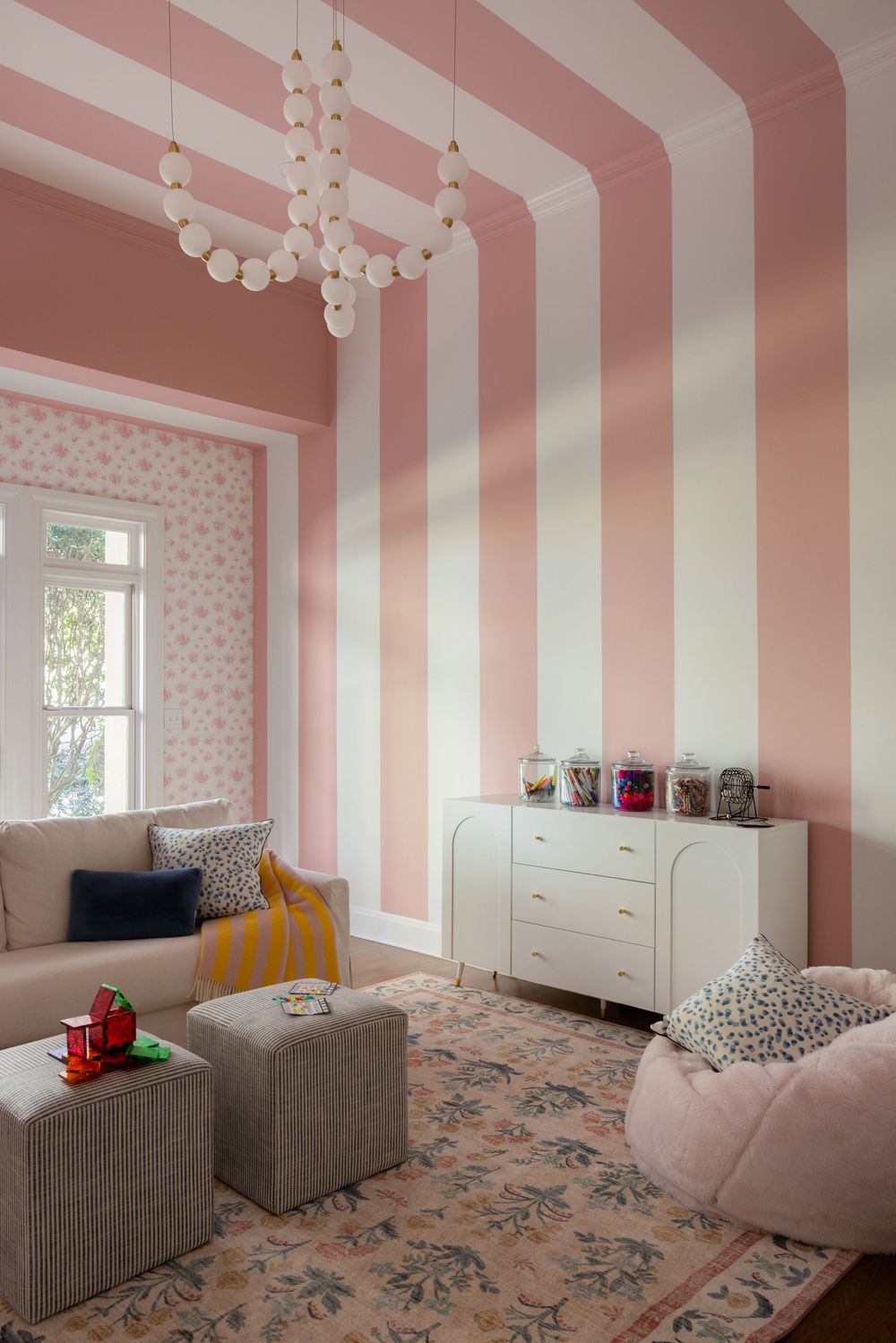 Pink striped nursery room with white dresser, globe chandelier, floral rug, and cozy seating in a soft pastel color scheme.