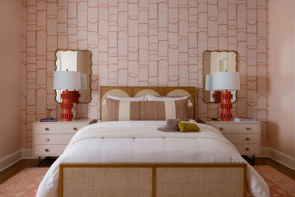 Elegant bedroom with pink geometric wallpaper, gold-framed mirrors, orange ceramic lamps, and a neutral upholstered bed with white bedding.