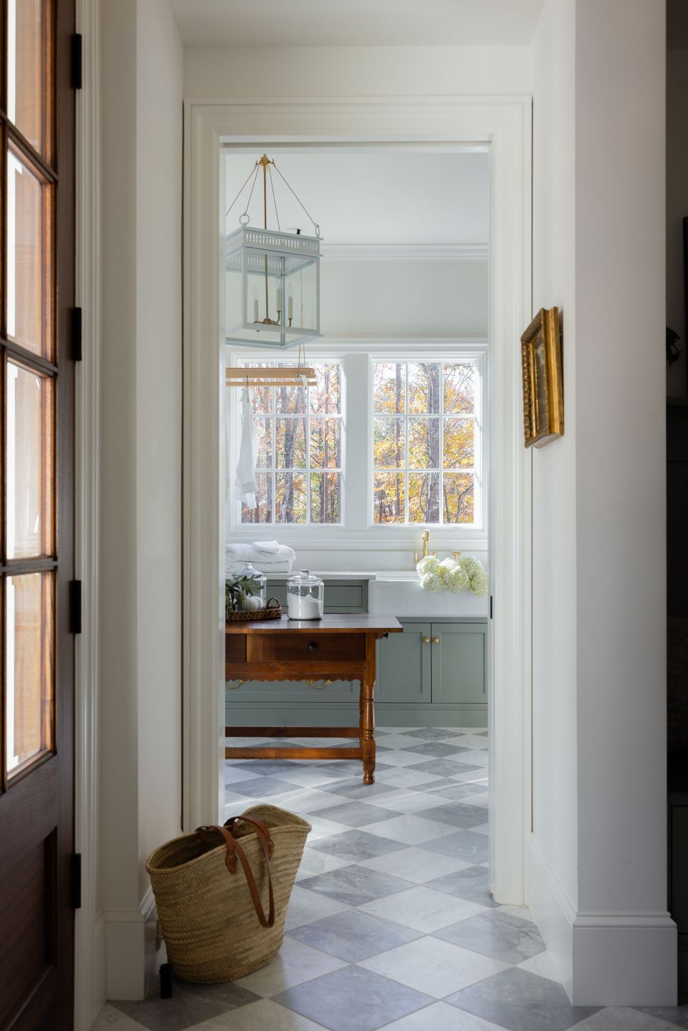 Elegant hallway with checkered marble floors leading to a bright laundry room with sage cabinets and brass lantern pendant.