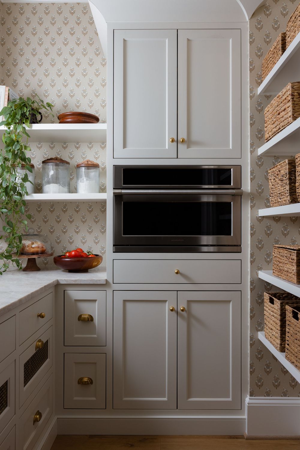 Gray shaker kitchen cabinets with brass hardware, built-in appliances, open shelving, and floral wallpaper in a stylish pantry.