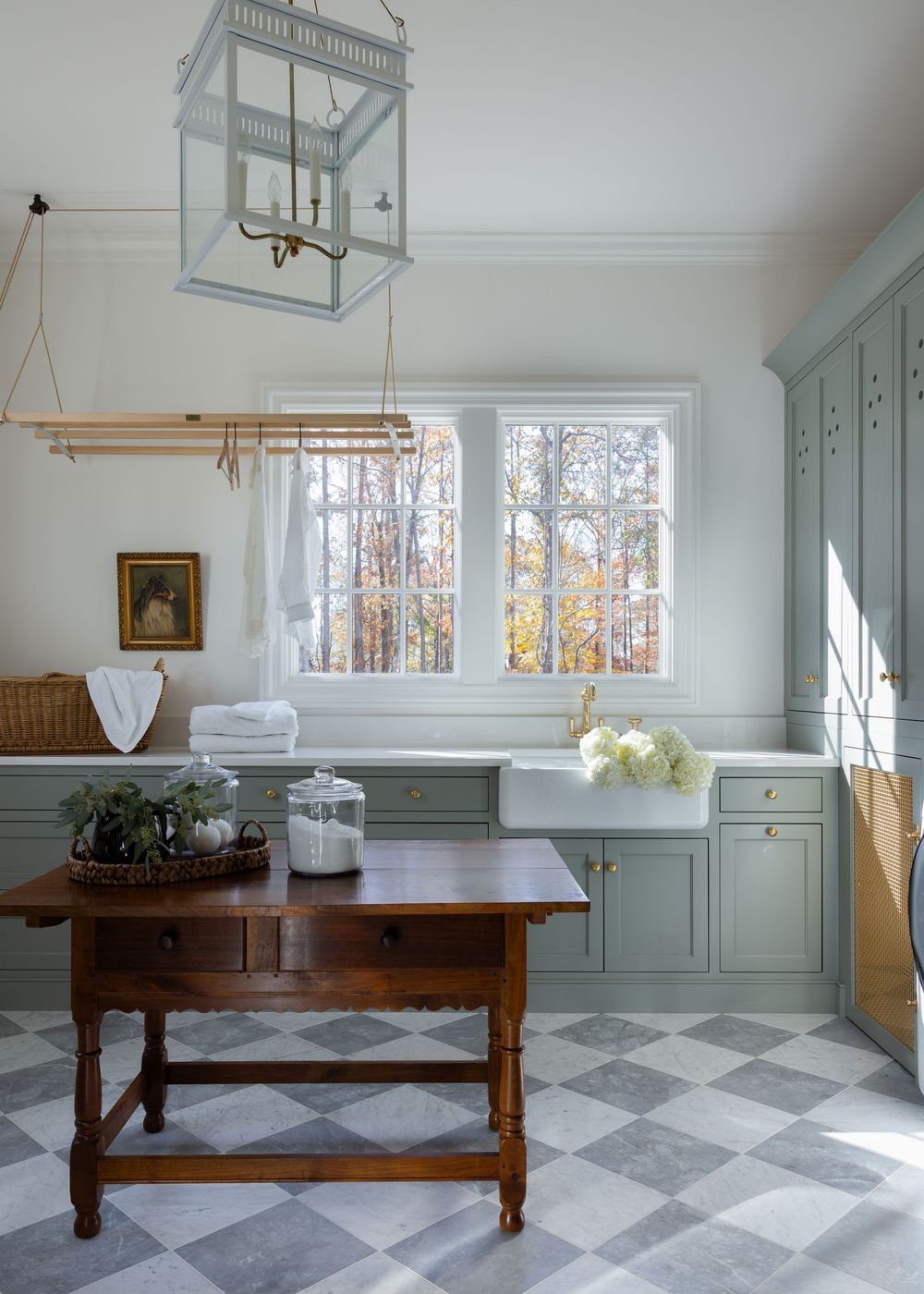 Elegant laundry room with sage green cabinets, checkered tile floor, antique wood table, farmhouse sink, and hanging drying rack.