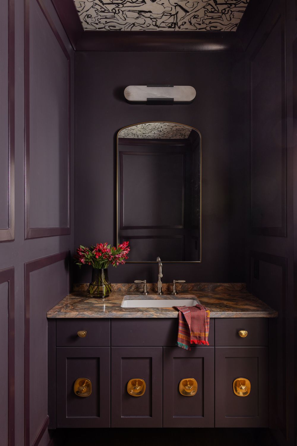 Elegant powder room with deep plum walls, marble vanity, gold hardware, arched mirror, and patterned ceiling.