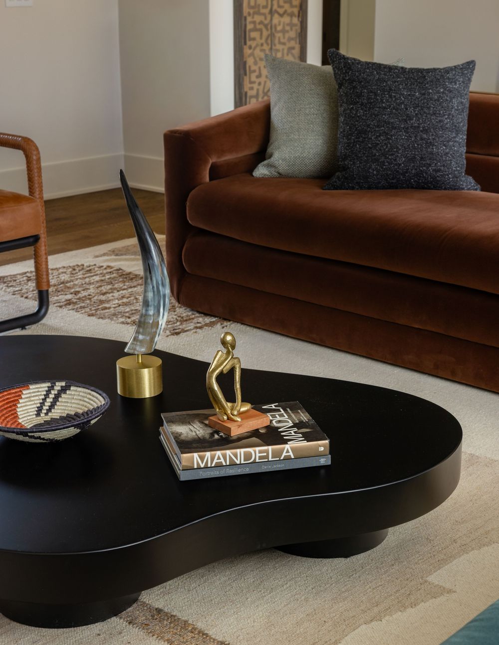 Black kidney-shaped coffee table styled with a Mandela book, gold figurine, and horn sculpture, set before a rust velvet sofa.