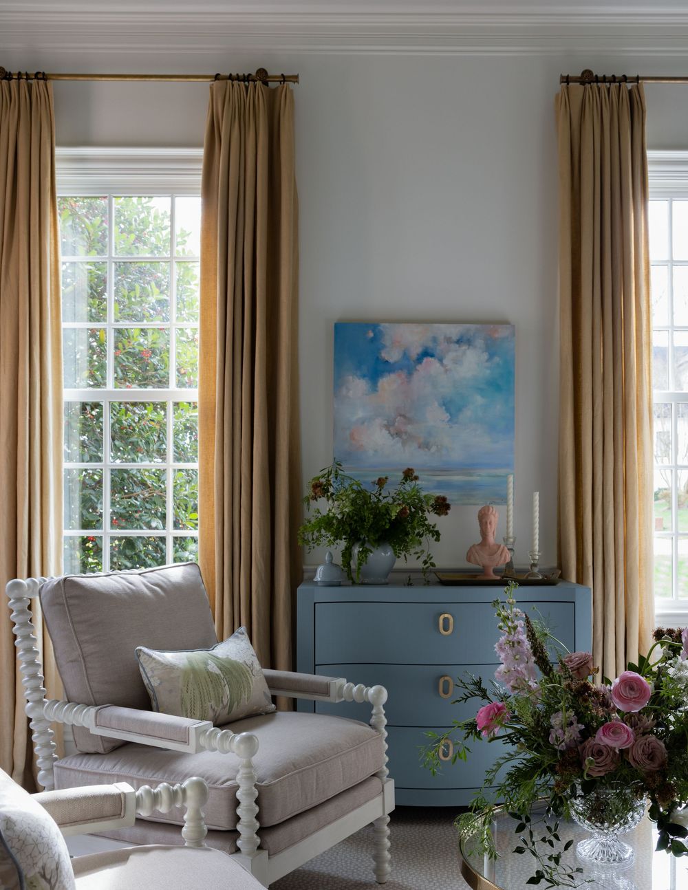 Elegant living room with gold curtains, blue dresser, cloud painting, and spindle armchair with floral arrangements.