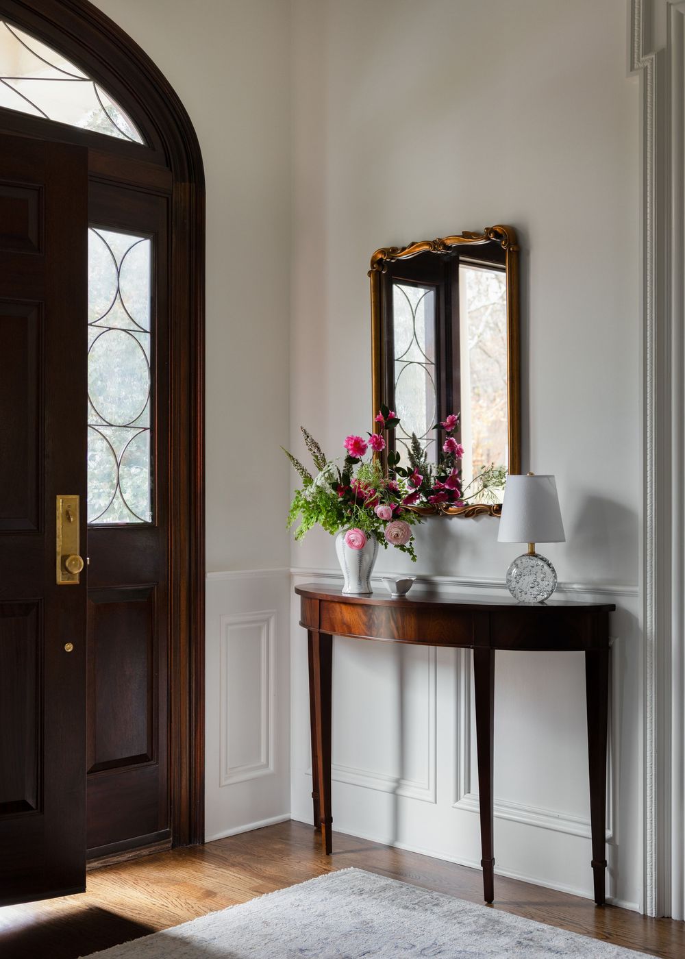 Classic entryway with dark wood arched door, demilune console table, gold mirror, and fresh floral arrangement.