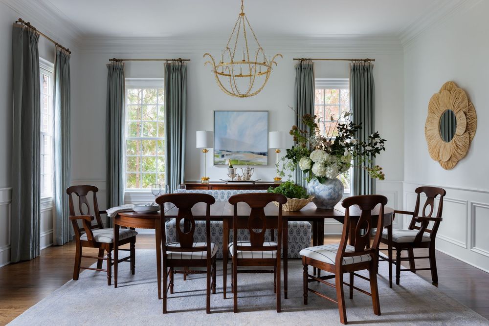 Elegant dining room with wooden chairs, oval table, gold chandelier, sage curtains, and soft blue-gray walls.