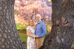 A couple stands between two trees with pink cherry blossoms blooming in the background during spring. Ottawa Wedding Photographer