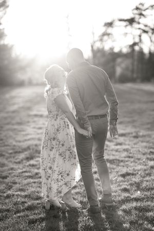 A romantic couple in a floral dress and dress shirt stands in a sunlit meadow with sun flare behind them. Ottawa Wedding Photographer
