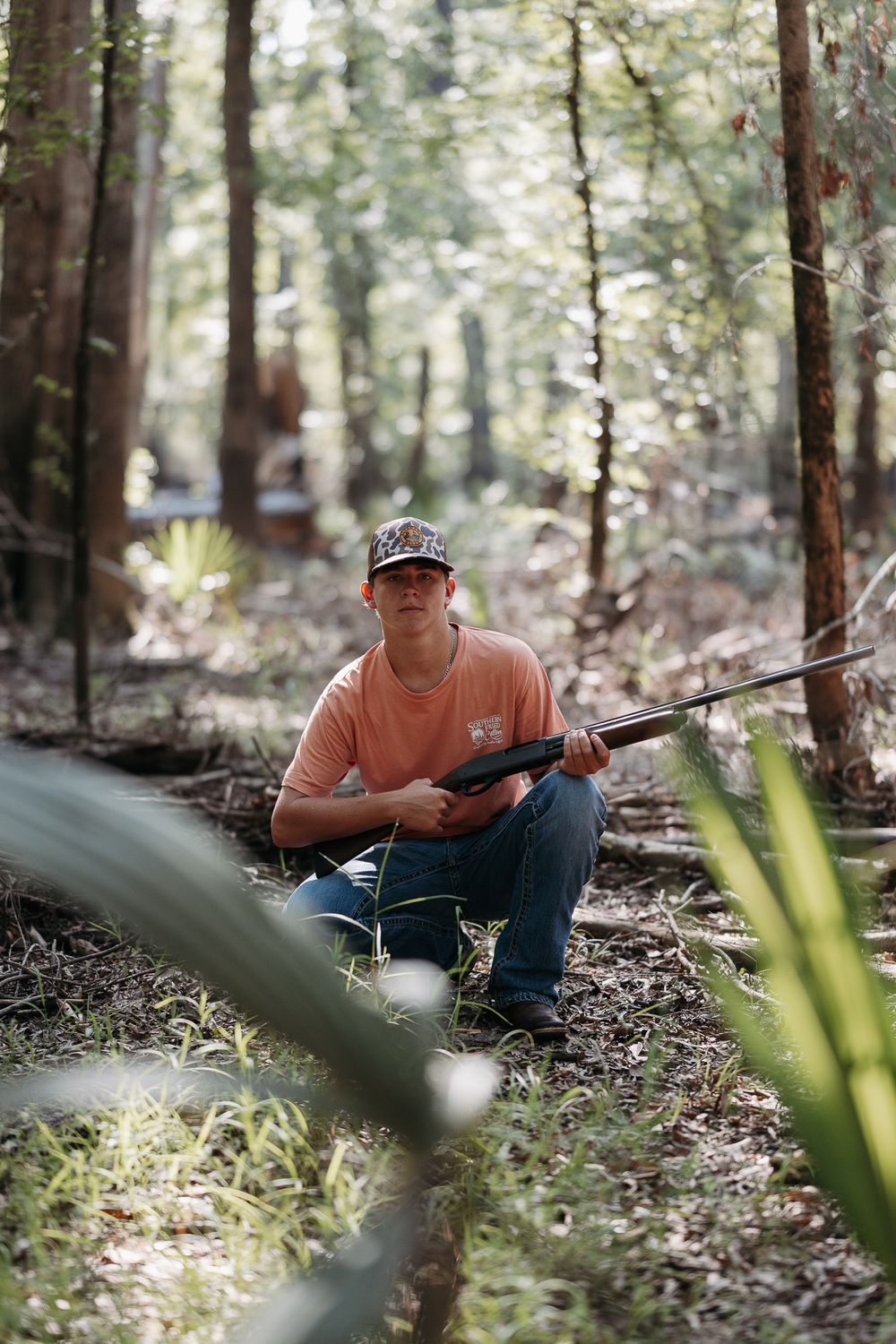Person in orange shirt sits on forest floor with camera equipment in natural woodland setting.