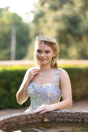 A formal portrait of someone wearing a delicate tiara and light blue embellished dress in an outdoor garden setting.