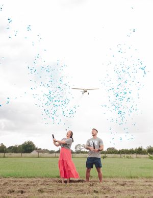 A couple releases blue confetti while a small airplane flies overhead in a grassy field.