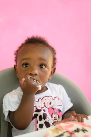 A toddler in a Minnie Mouse shirt with cake frosting on their face against a pink backdrop.