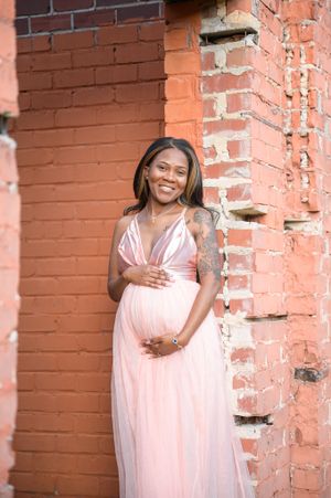 Pregnant woman in pink dress smiling against rustic brick wall.