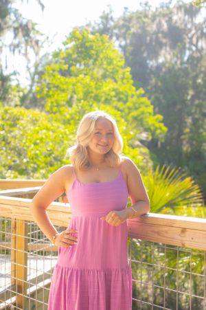 Woman in pink sundress poses on wooden deck with tropical foliage.