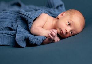 A newborn baby wrapped in blue fabric gazes at the camera during a professional photoshoot.