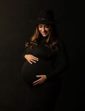 A maternity portrait of a pregnant woman in black wearing a wide-brimmed hat against dark background.