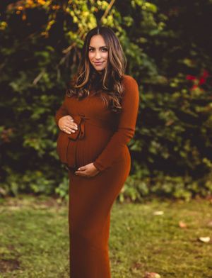 An expectant mother in a brown dress poses outdoors against a leafy background.