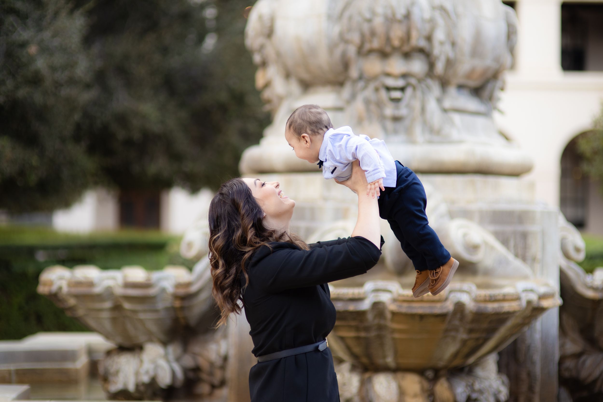 Parent lifting baby in front of ornate fountain in outdoor garden setting.