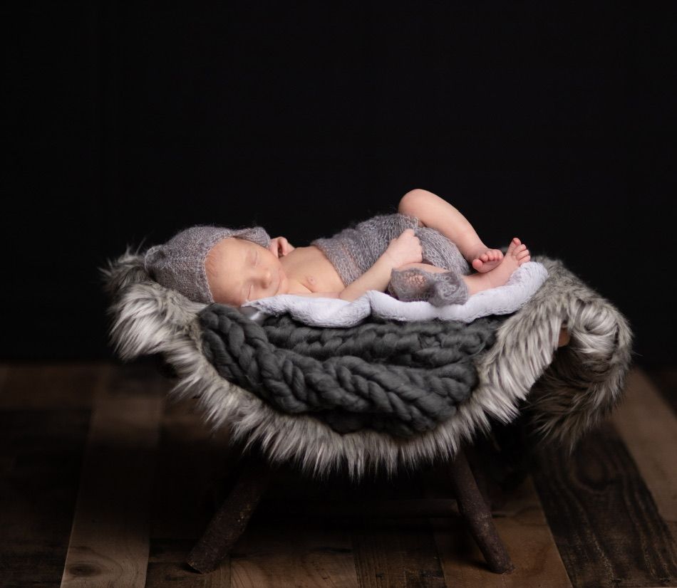 A newborn baby sleeps on a furry grey backdrop during a professional photoshoot.