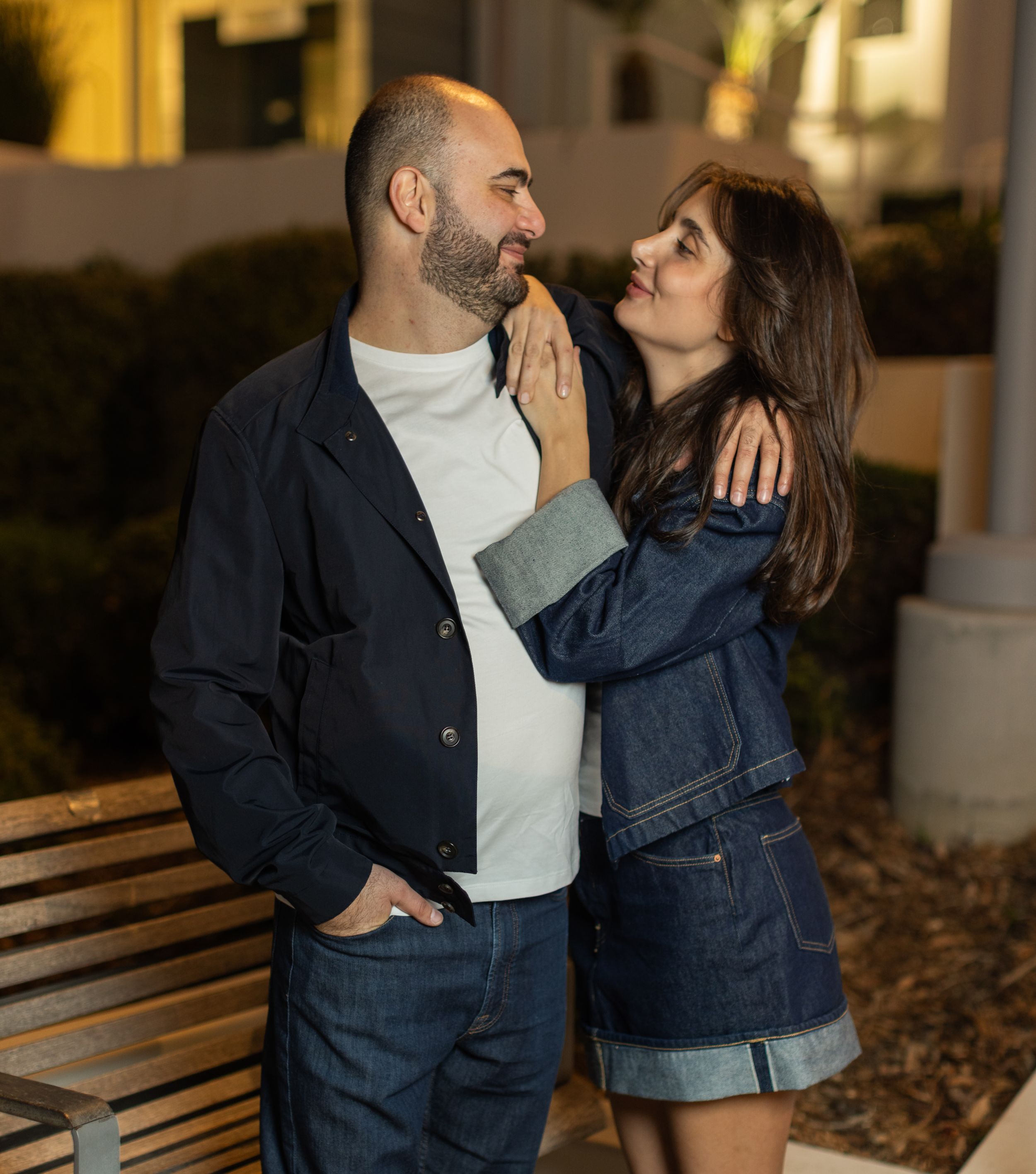 Couple in casual denim outfits sharing intimate moment on bench at dusk.