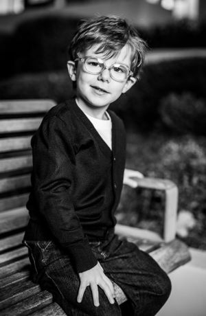 Black and white portrait of young child wearing glasses sitting on bench.