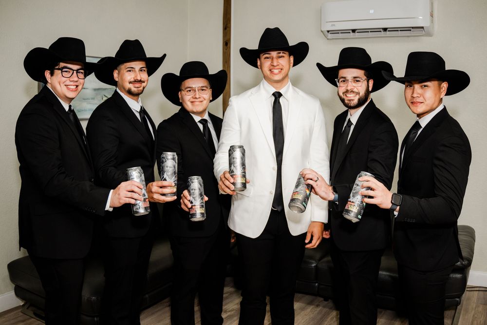 Six men in black suits and cowboy hats pose with canned drinks, one wearing a white jacket, at a formal event.