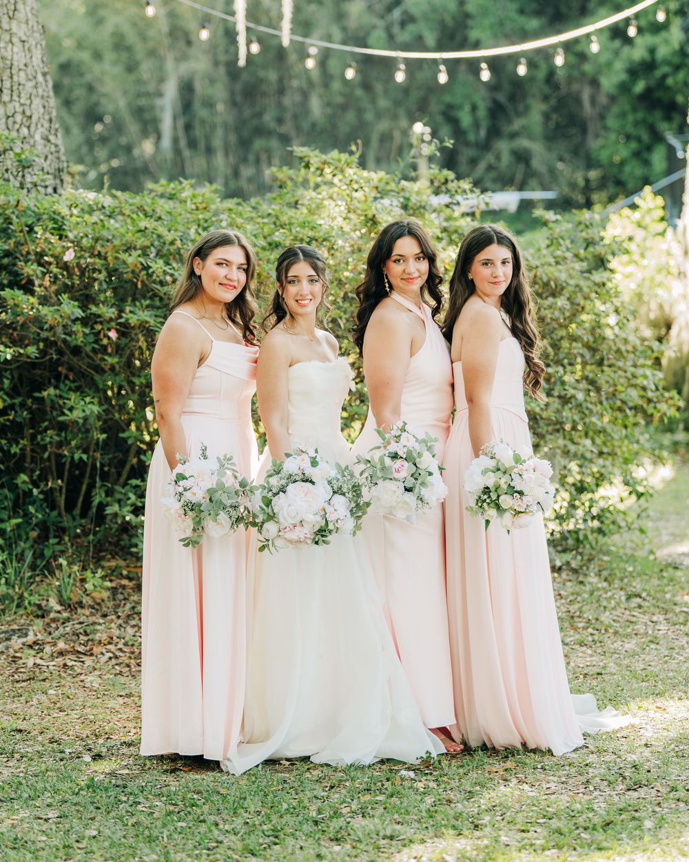Bride in white gown with three bridesmaids in blush pink dresses holding floral bouquets outdoors.