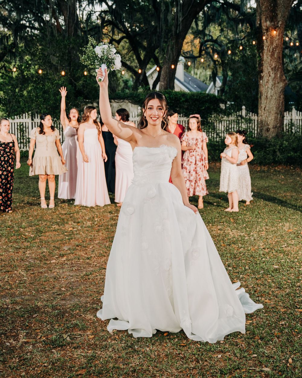 Bride in white gown tossing bouquet outdoors at wedding reception with guests watching in background.