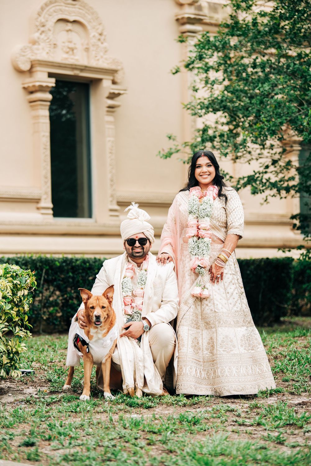 Indian couple in traditional wedding attire pose with their dog outside a ornate building.