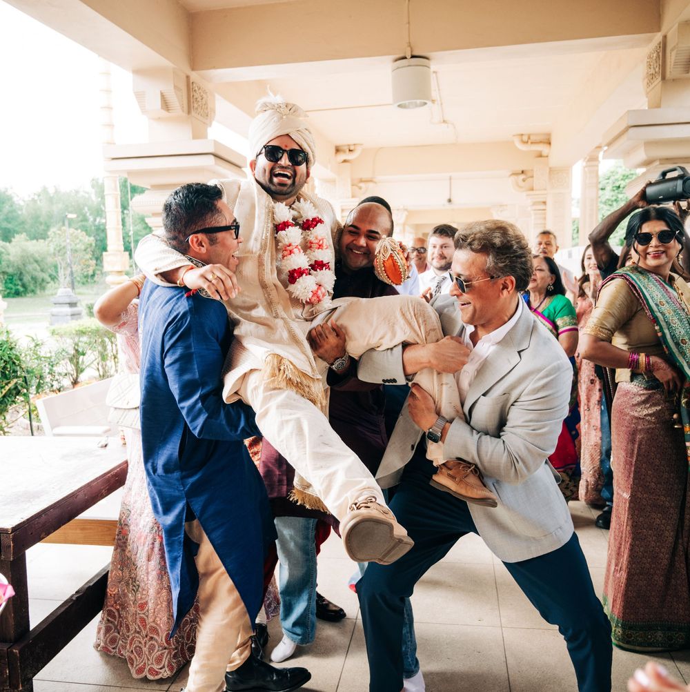 Groom in white sherwani and turban being lifted by groomsmen during joyful Indian wedding celebration outdoors.
