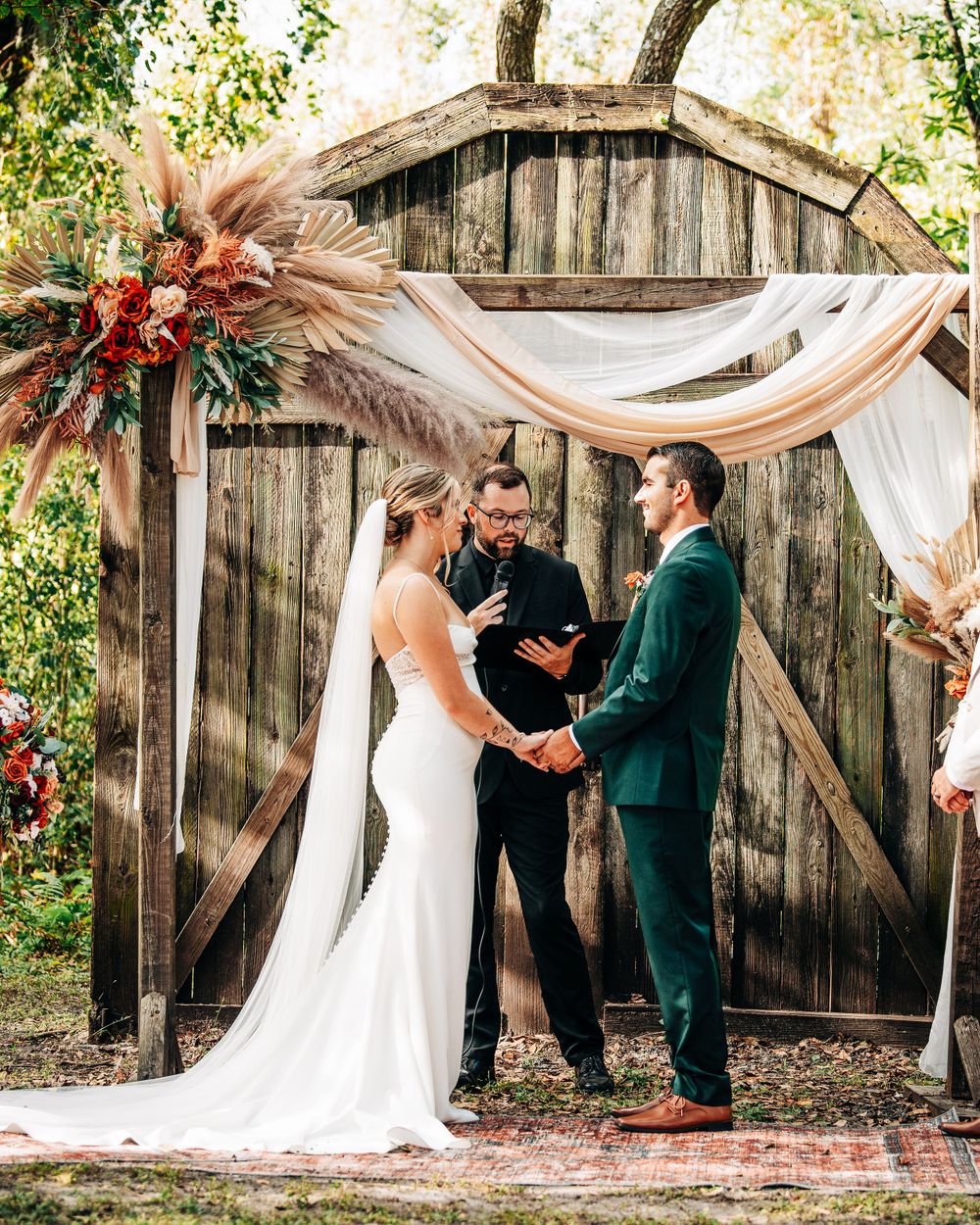 An outdoor wedding ceremony under a rustic wooden archway decorated with flowy white fabric and pampas grass.