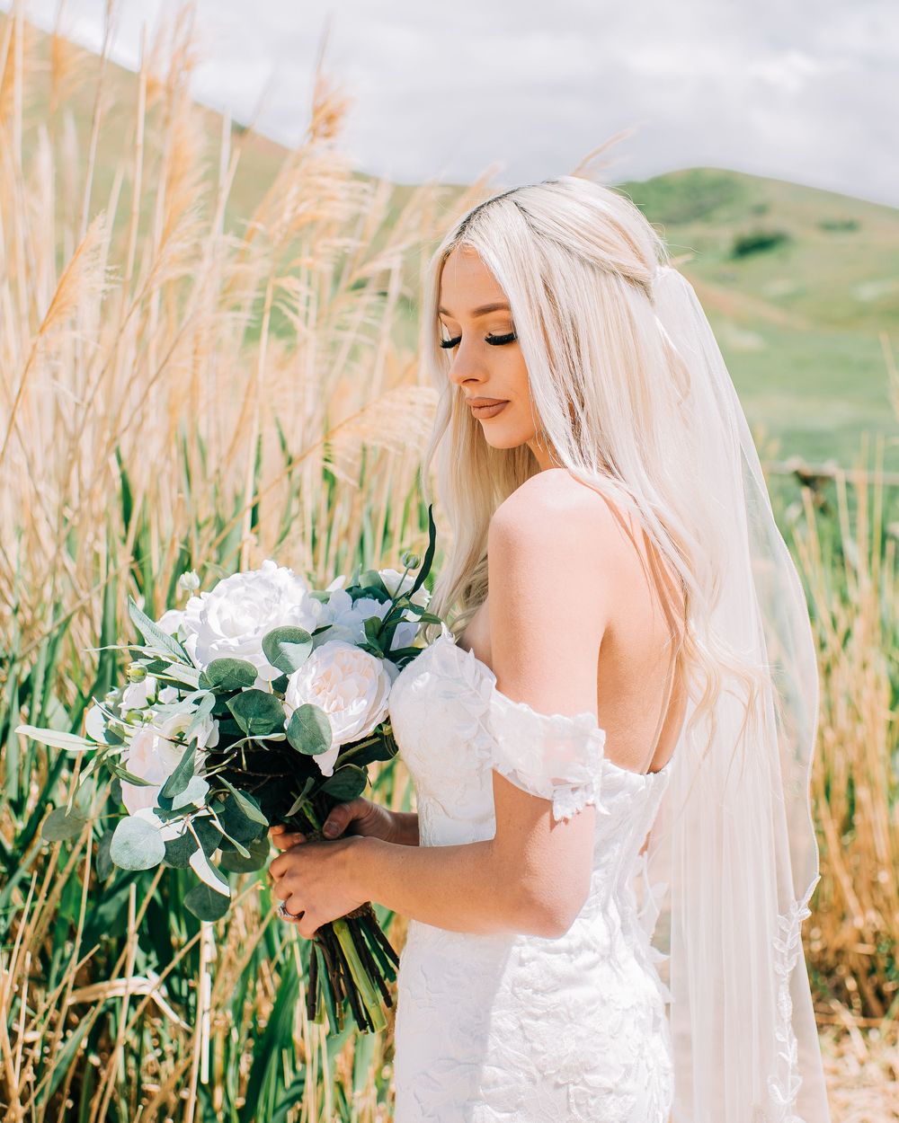 Blonde bride in white lace off-shoulder gown holds white floral bouquet with greenery in golden field.