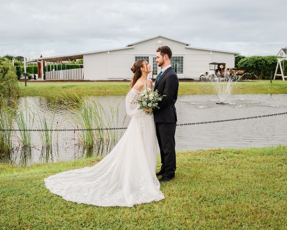 Bride and groom pose by a serene pond with a fountain and white barn venue in the background on their wedding day.
