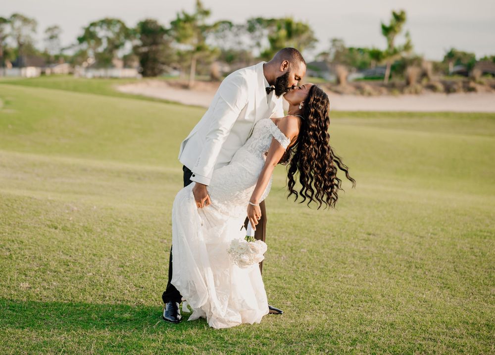 A groom in a white tuxedo dips his bride in a white gown for a romantic kiss on a lush green golf course.