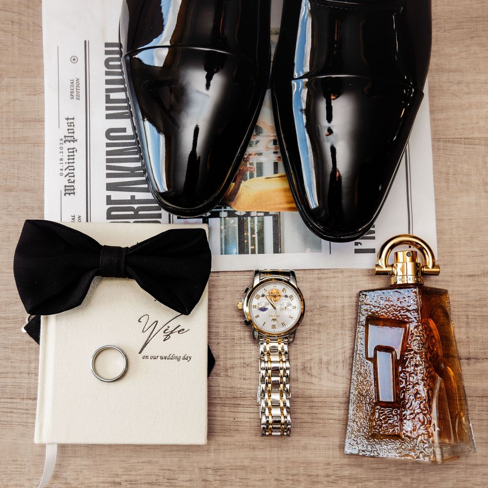 Groom wedding flat lay with black patent shoes, bow tie, watch, cologne, ring, and Wife card on wood.