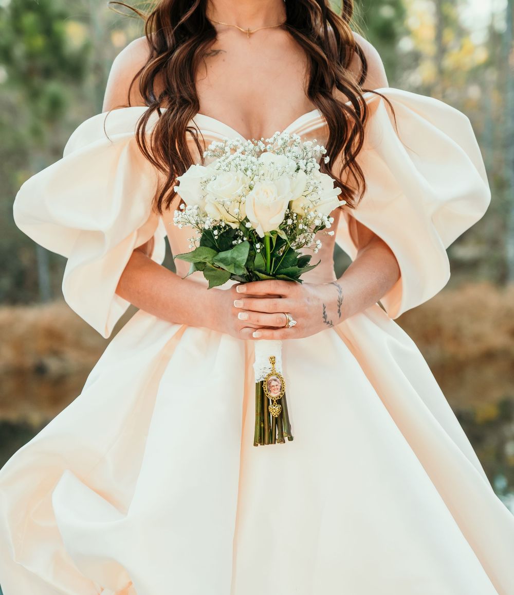 Bride in off-shoulder blush ball gown holding white rose bouquet with gold tassel charm at outdoor wedding.