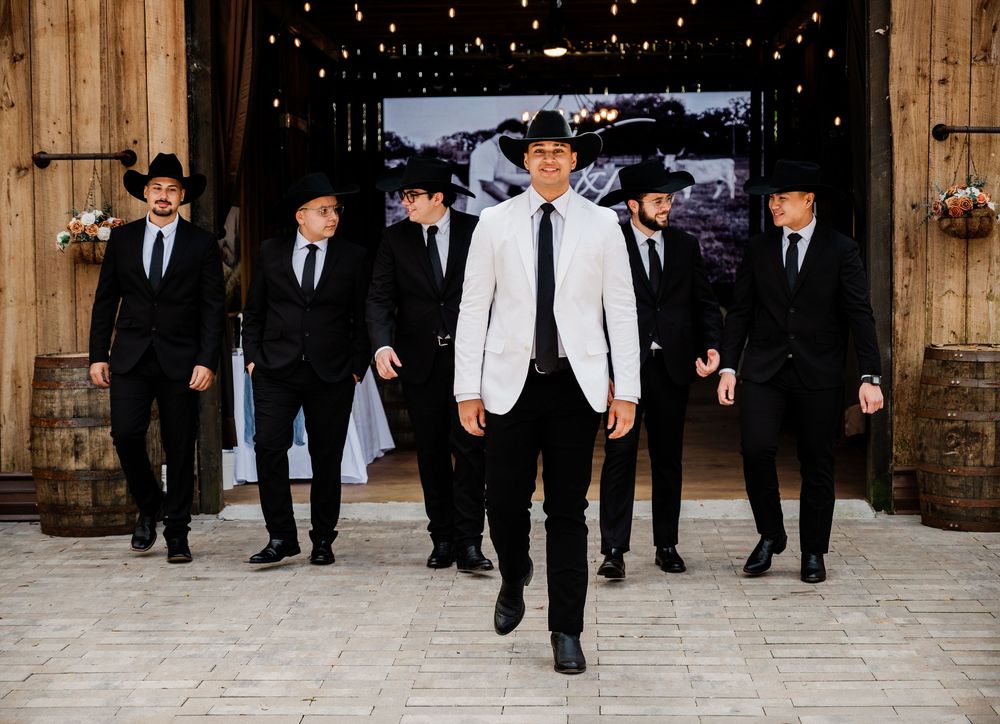 Groom in white blazer leads groomsmen in black suits and cowboy hats through rustic barn wedding venue.