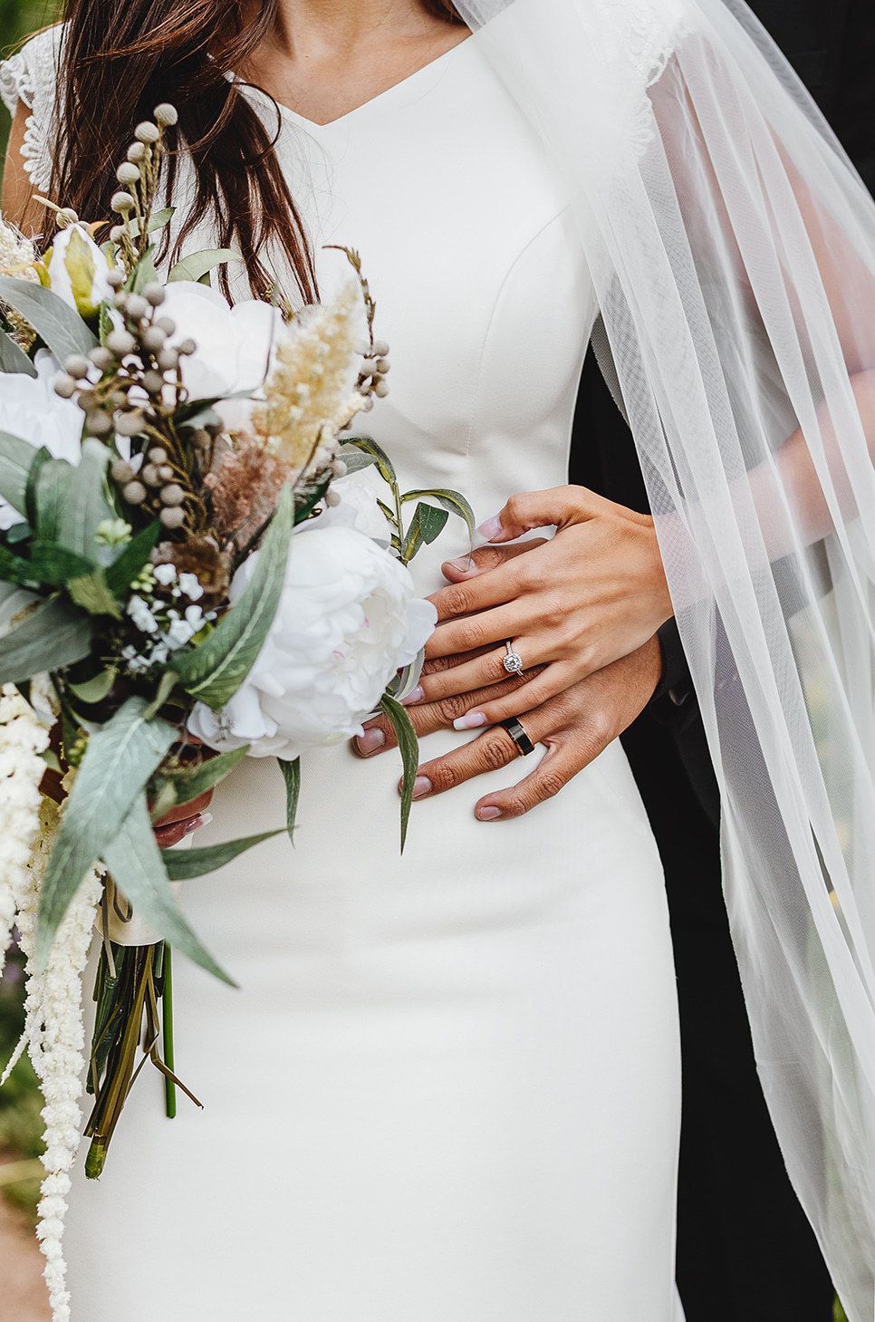 Elegant white wedding dress with flowing veil and cascading wildflower bouquet showing engagement ring detail.
