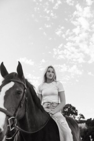 Black and white photo of a rider atop a dark horse in casual attire against a cloudy sky.